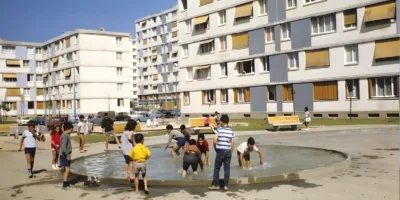 Des enfants jouent joyeusement dans un bassin d'eau public, devant des immeubles d'habitation.