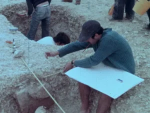 Archéologue mesurant un artefact dans une fouille, ambiance rétro et terrain rocailleux.