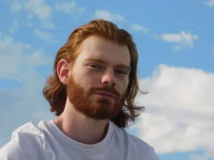 Un jeune homme roux avec une barbe et des longs cheveux, souriant face au ciel bleu.