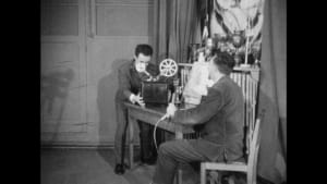 Deux hommes utilisent un projecteur de cinéma dans un décor d'époque.