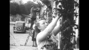 Des femmes récoltent des raisins sur un treillis dans un village, en noir et blanc.