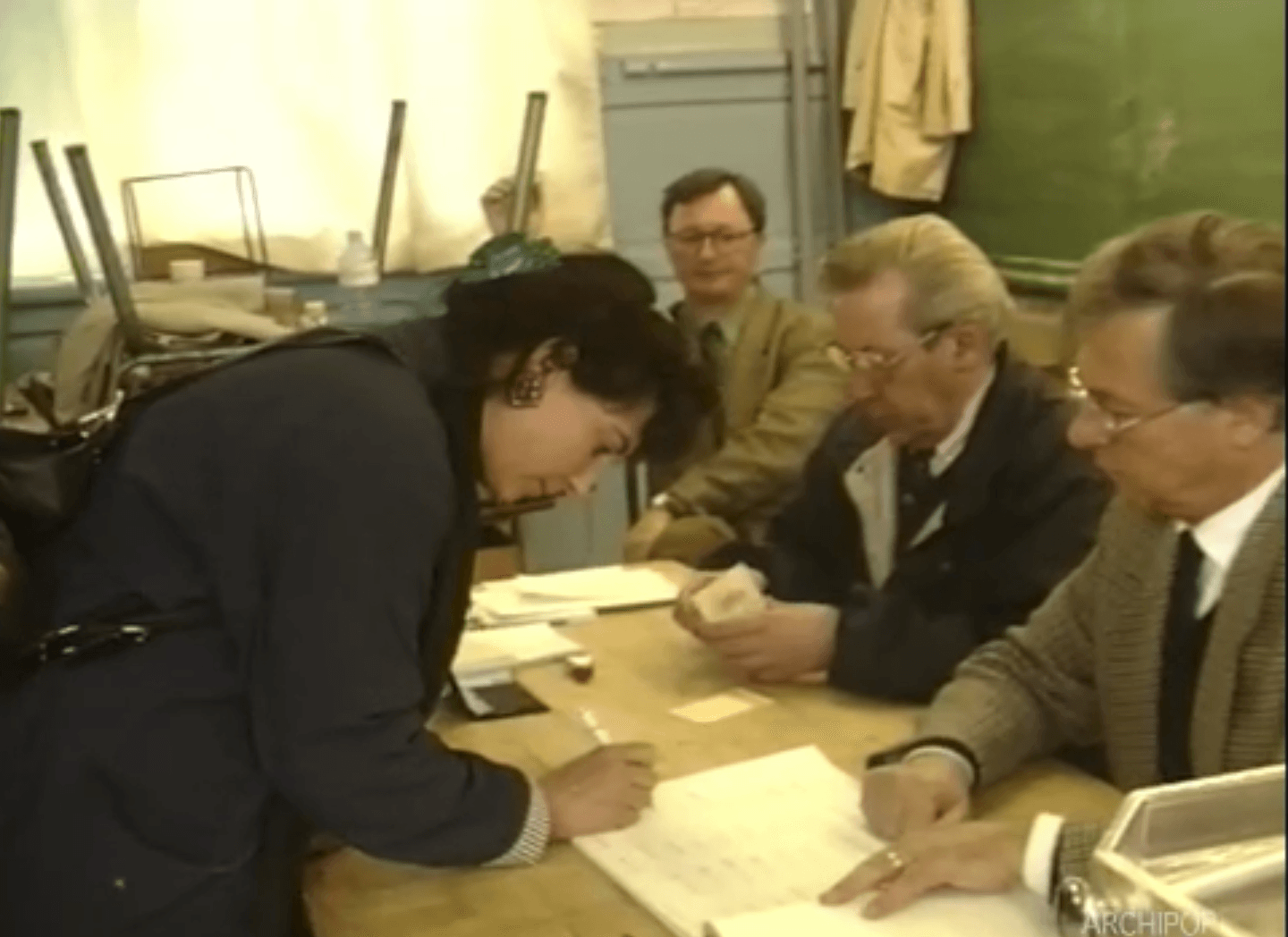 Une femme signe des documents avec l'aide de collègues dans un bureau des années 80.