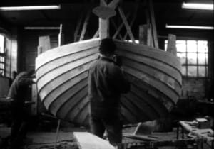 Photographie en noir et blanc de deux charpentiers de marine construisant la coque d'un bateau en bois.