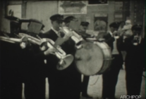 Une fanfare d'hommes en uniformes sombres jouant de divers instruments à vent et d'un tambour.
