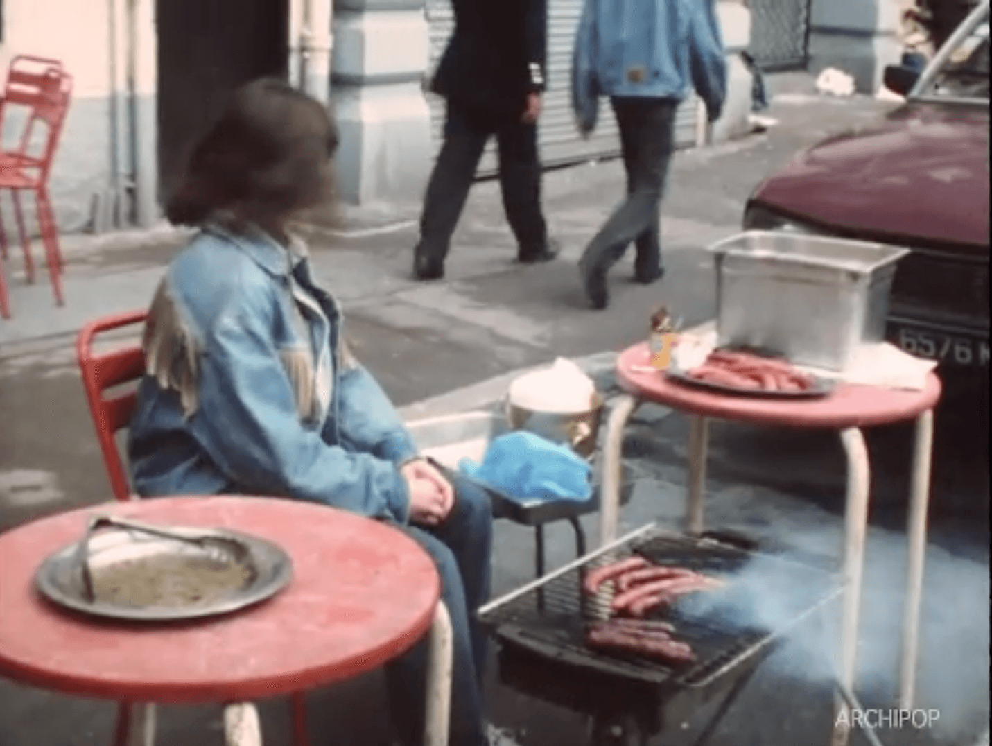 Une femme en veste bleue est assise à une table rouge à côté d'un grill avec des saucisses.