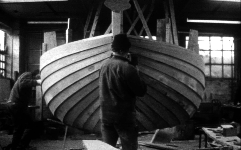 Photographie en noir et blanc de deux charpentiers de marine construisant la coque d'un bateau en bois.