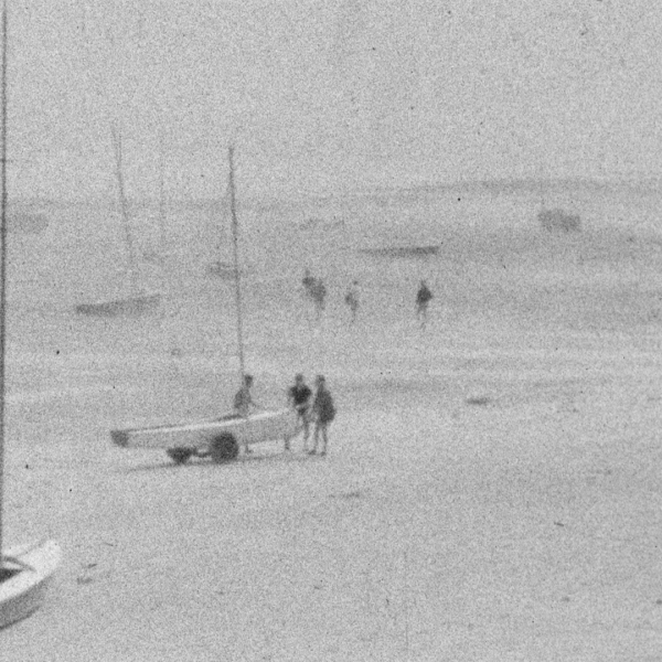 Photographie ancienne en noir et blanc de voiliers et de personnes sur une plage brumeuse.