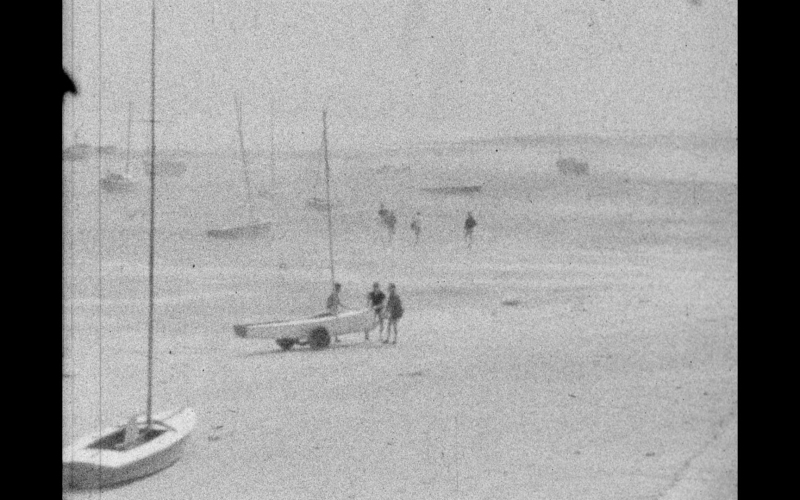 Photographie ancienne en noir et blanc de voiliers et de personnes sur une plage brumeuse.
