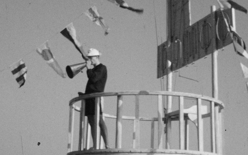 Un vigile maritime utilise une mégaphone depuis une tour de guet, entouré de drapeaux.