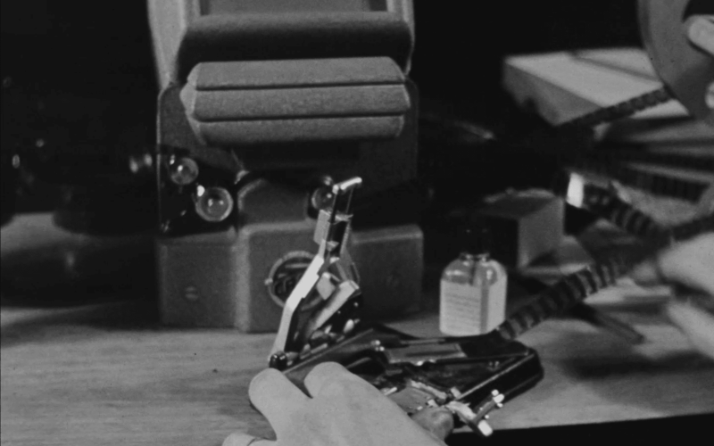 Une main manipule un outil métallique sur un bureau encombré d'appareils électroniques anciens en noir et blanc.