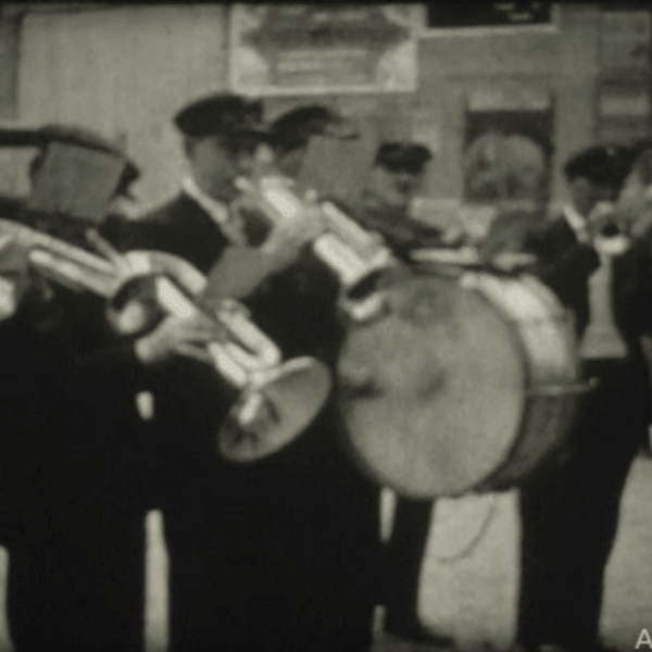Une fanfare d'hommes en uniformes sombres jouant de divers instruments à vent et d'un tambour.