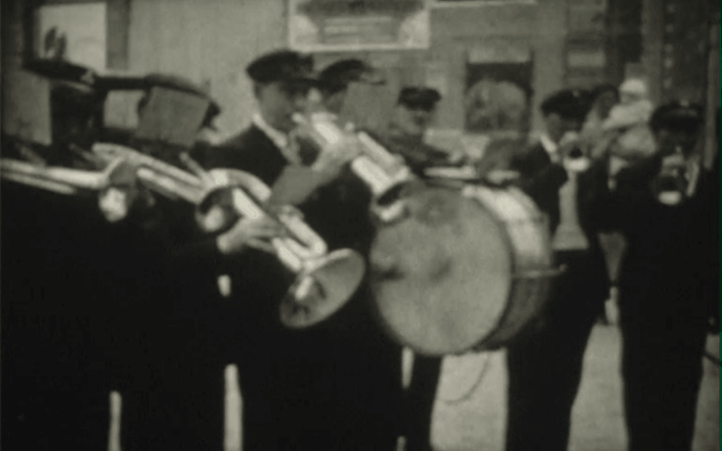 Une fanfare d'hommes en uniformes sombres jouant de divers instruments à vent et d'un tambour.