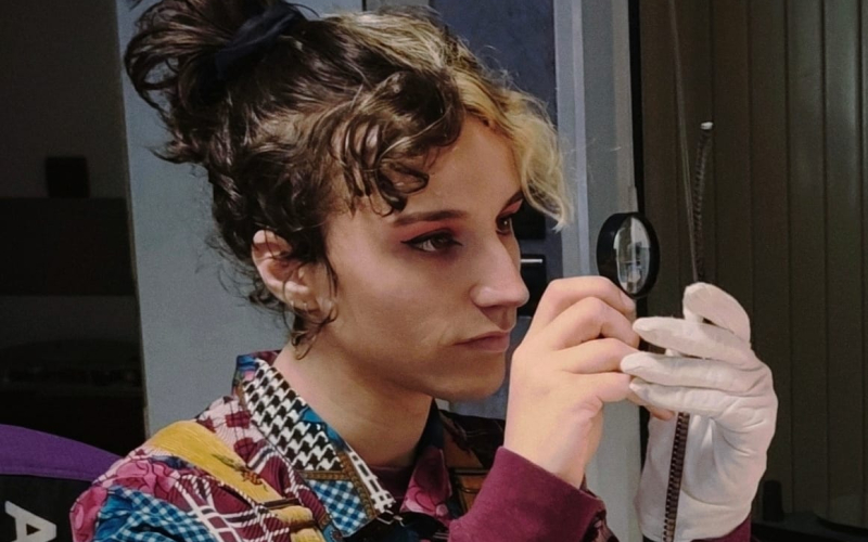 Une personne aux cheveux bicolores et maquillage marqué regarde à travers une loupe.