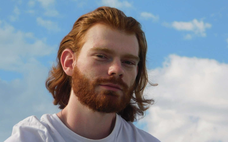 Un jeune homme roux avec une barbe et des longs cheveux, souriant face au ciel bleu.